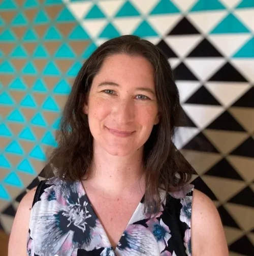 A woman with shoulder-length brown hair, smiling, wearing a sleeveless floral top, standing in front of a geometric background with blue, black, and white triangles.