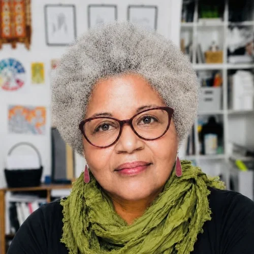 Portrait of an older woman with gray curly hair wearing glasses, a green scarf, and pink earrings, inside a room with shelves and artwork in the background.