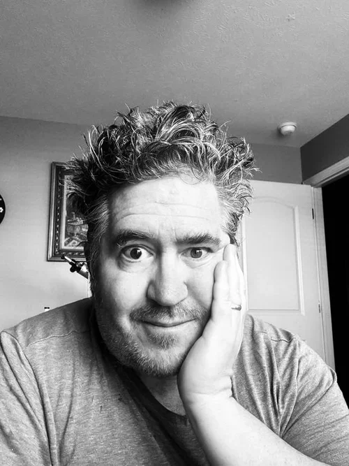 Black-and-white photo of a man with messy hair, resting his face on his hand and smiling, indoors with a picture frame on the wall behind him.