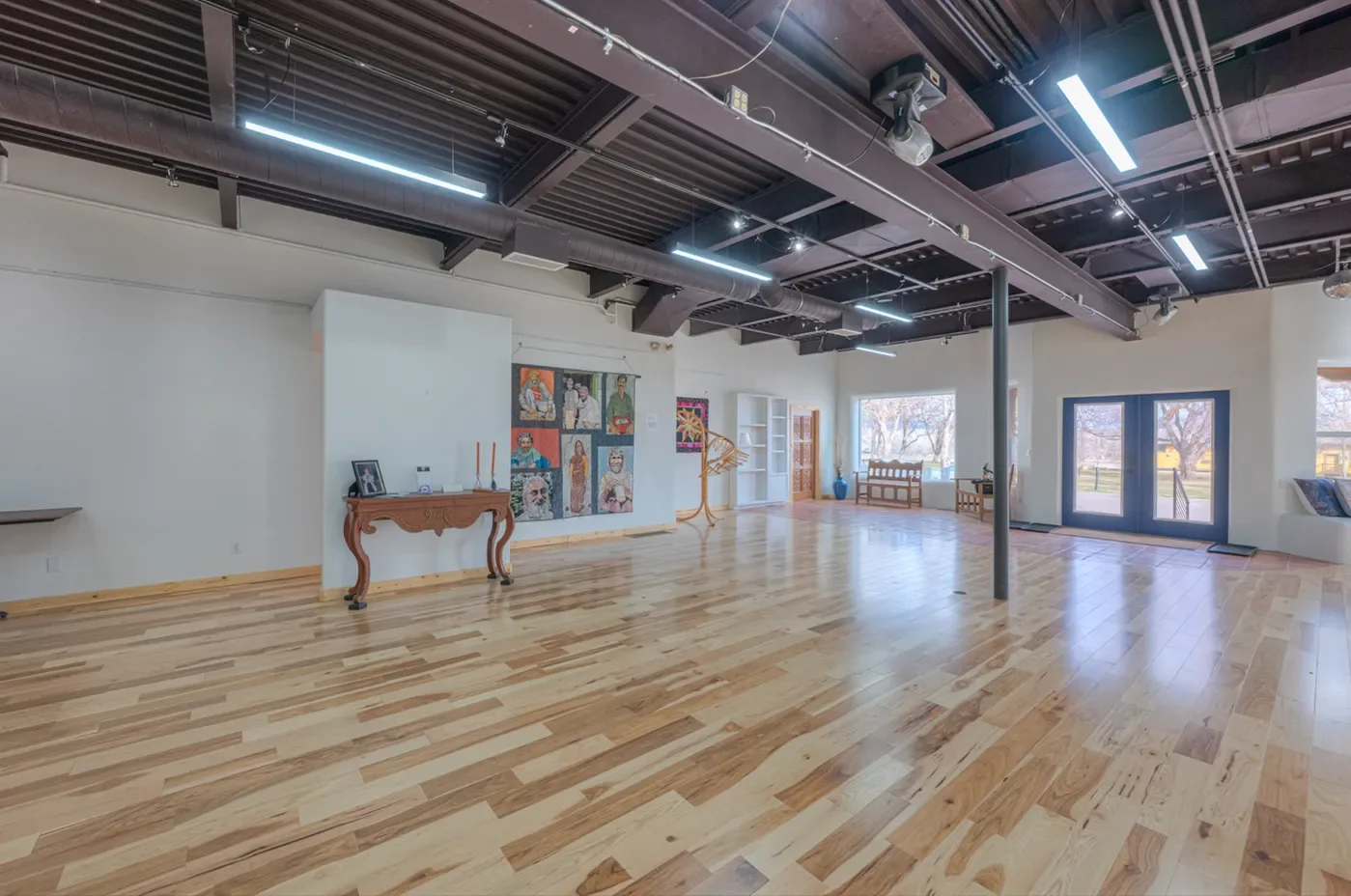 An empty art gallery with wooden floors, white walls, and large windows. There are artworks on the walls, a couple of benches, and display shelves. The ceiling has exposed black beams and track lighting.