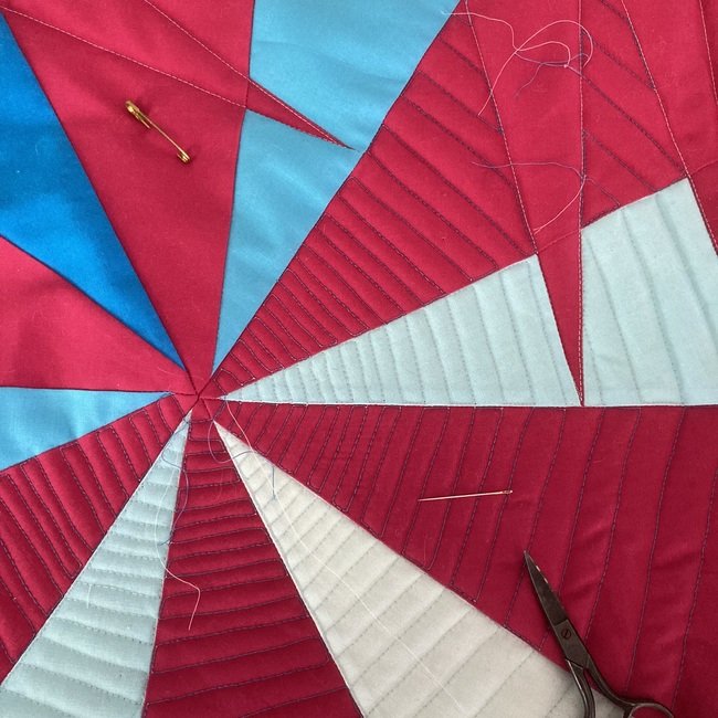 Close-up of a multicolored quilt featuring red, light blue, dark blue, and white fabric sections, with sewing needles and scissors on the quilt.