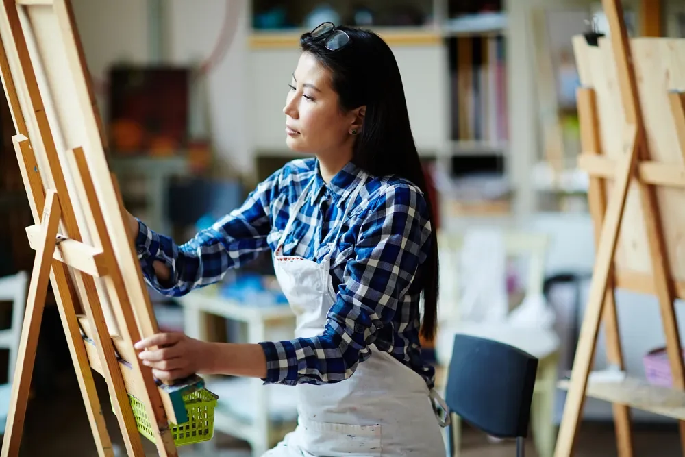 A woman in a checkered shirt and apron painting on a canvas using an easel in an art studio.