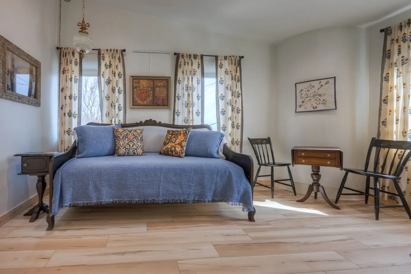 A vintage living room with a sofa covered by a blue throw, three decorative pillows, three windows with patterned curtains, a small wooden table, two black chairs, wall art, and a wooden floor.