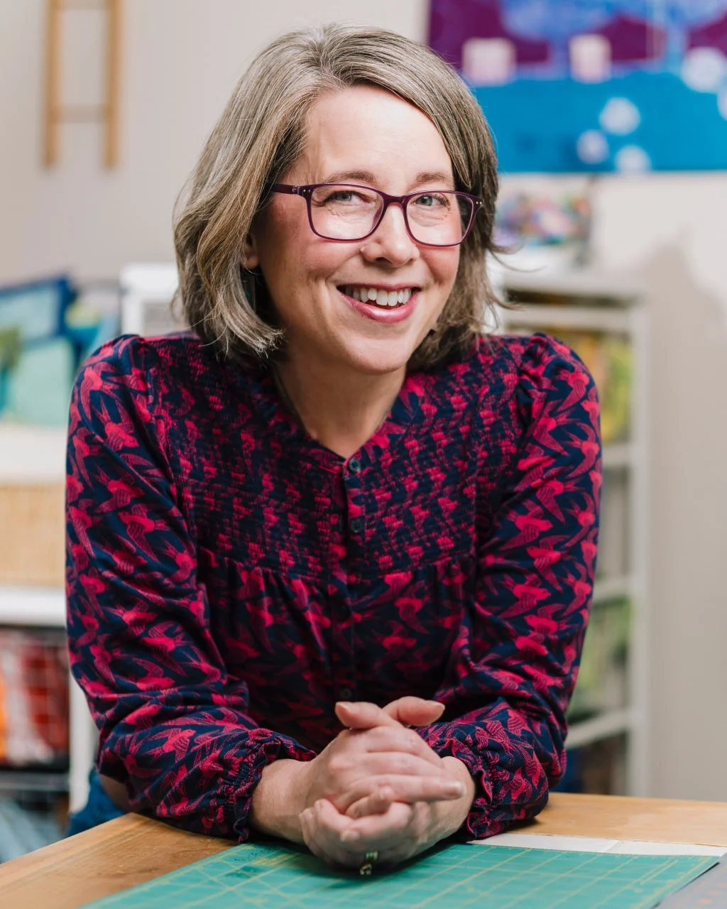 A woman with shoulder-length gray hair, glasses, and a red and blue patterned blouse sitting at a table in a classroom or craft room, smiling at the camera.