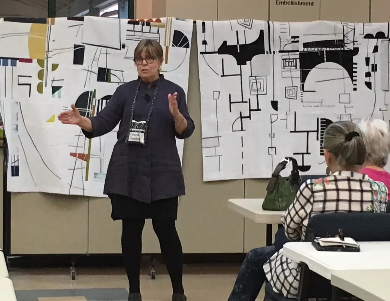 A woman with glasses and a dark jacket is giving a presentation in front of large abstract black and white artwork and talking to an audience of women seated at tables in a conference room.
