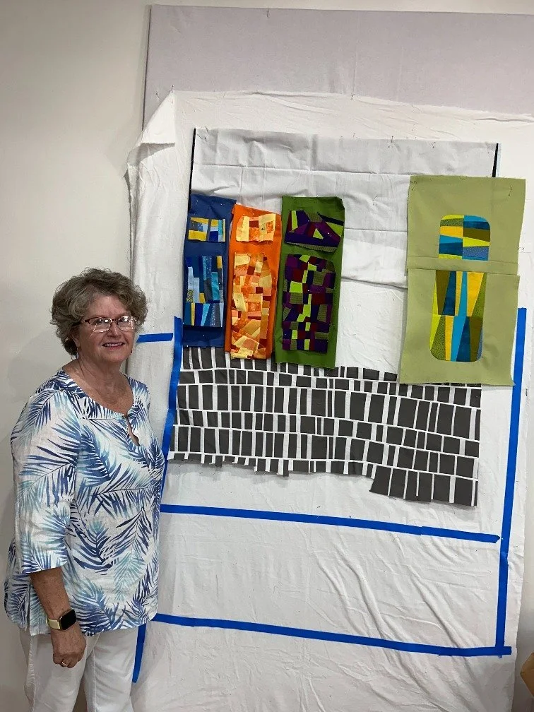 A woman with curly gray hair, glasses, and a white and blue patterned shirt stands smiling next to a fabric art piece or quilt with colorful vertical rectangular shapes and black and white striped patterns on a white background.