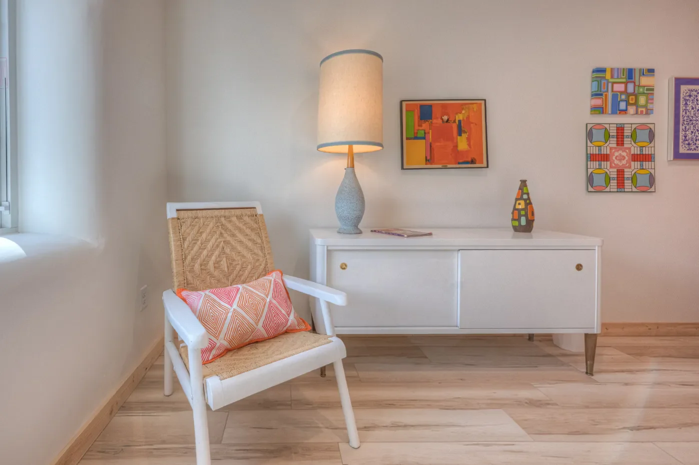 Living room corner with a white chair with woven back and a pink geometric pillow, a white cabinet with colorful vases and abstract art, a large table lamp, and framed artwork on the wall.