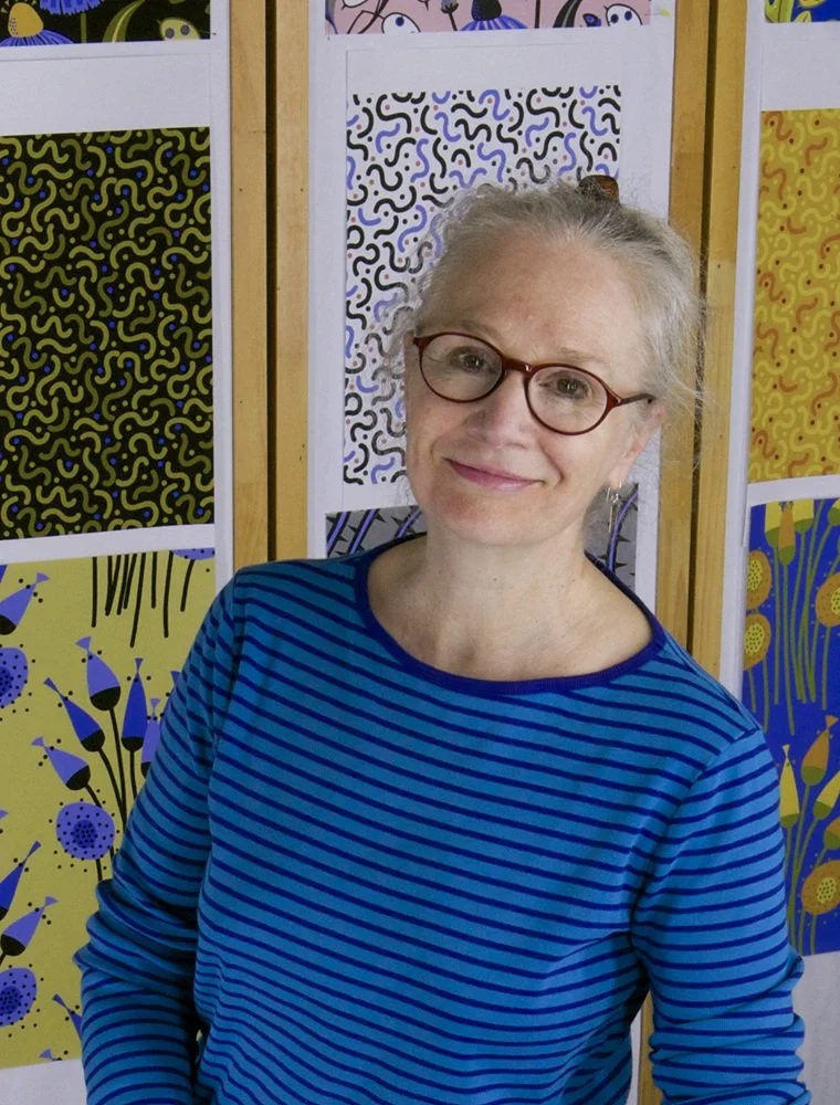 A woman with glasses and gray hair, wearing a blue and black striped shirt, standing in front of colorful abstract artwork displayed on a wooden partition.