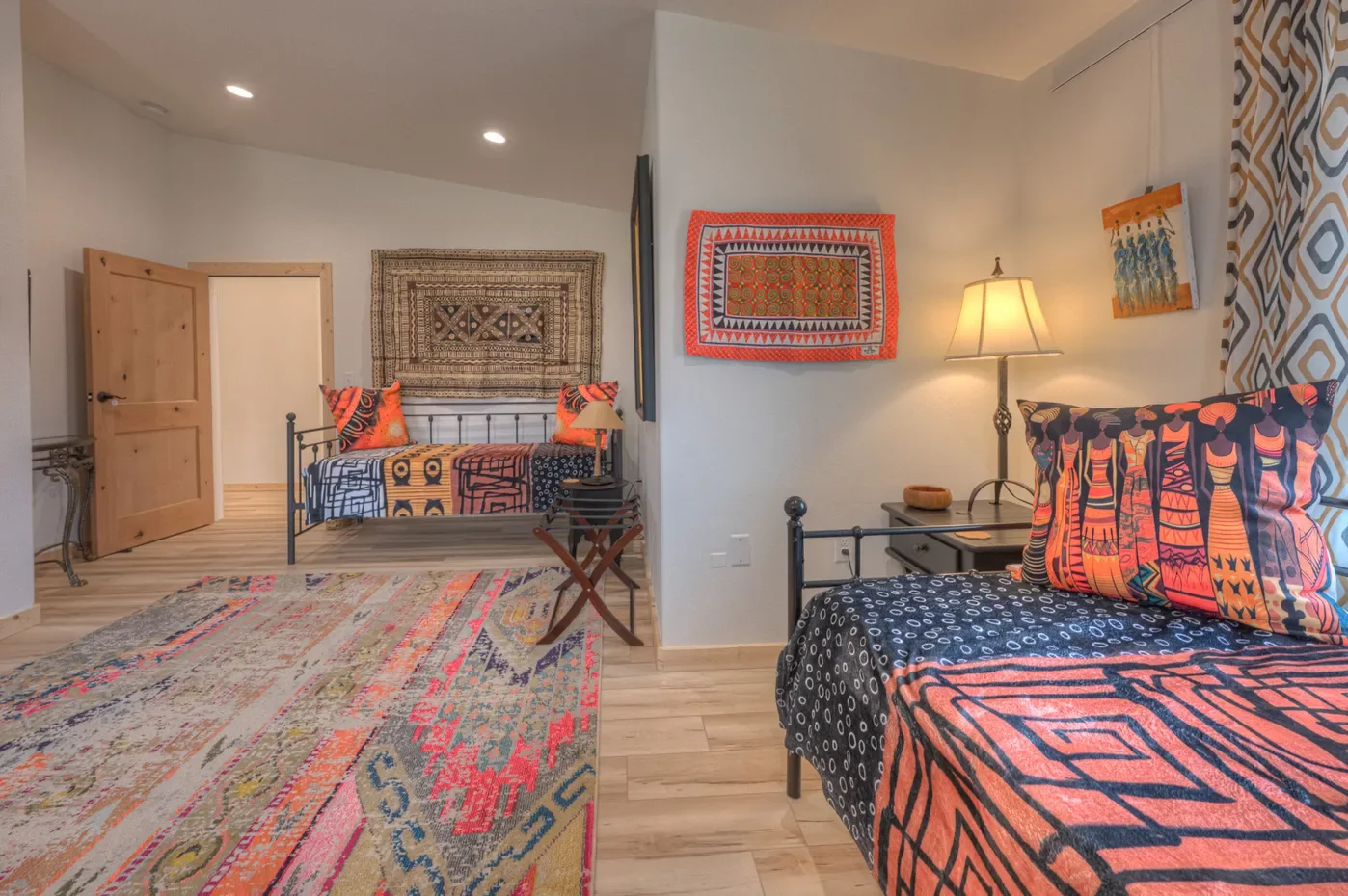 Decorative bedroom with two beds, colorful patterned textiles, wall art, lamp, and hardwood flooring.