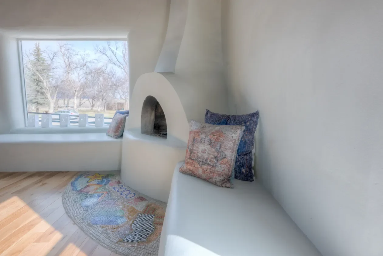 A cozy corner with a built-in white bench by a window, colorful pillows, and a small tiled fireplace, with a view of trees and parked cars outside.