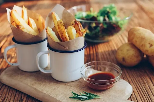 Two white mugs with French fries wrapped in paper, a small bowl of dipping sauce, and a bowl of salad with potatoes on a wooden cutting board.