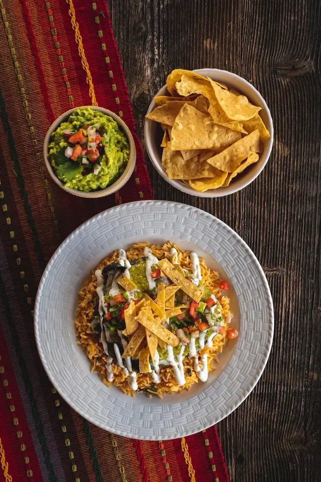 A plate of Mexican rice topped with sour cream, guacamole, chopped tomatoes, and crispy tortilla strips, with a side bowl of guacamole with diced tomatoes, and another bowl of tortilla chips on a rustic wooden table.