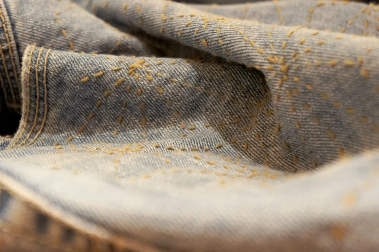 Close-up of blue denim fabric with yellow stitching and small yellow paint splatters.