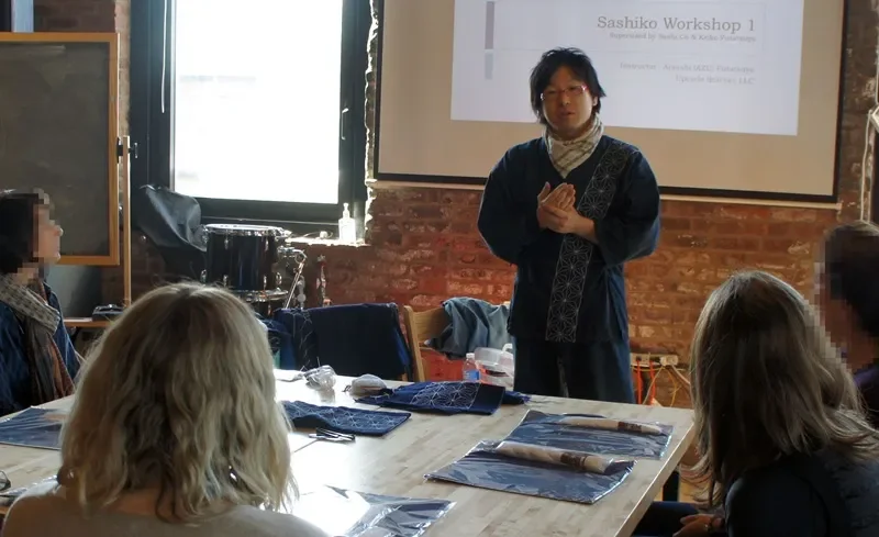 A man giving a presentation in a workshop, with a slide on the screen titled 'Sashiko Workshop 1', in a room with brick walls and a large window. Several women are seated around the table, listening attentively.
