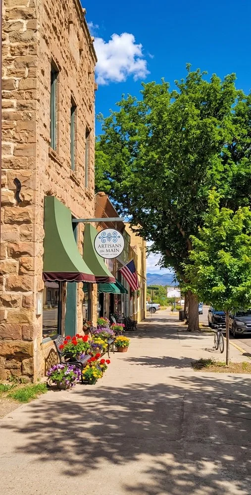 A street scene featuring a stone building with a green awning, colorful flower planters, a sidewalk with trees, parked cars, and a sign reading 'Artisans on Main' on a sunny day with a bright blue sky and some clouds.