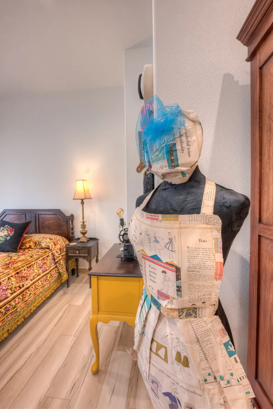 A dress form covered with newspaper-style fabric, wearing a matching apron and headwrap made of similar material, standing beside a yellow side table in a bedroom. The bedroom features a bed with vibrant floral bedding and a wooden headboard, a nightstand with a lamp, and hardwood floors.
