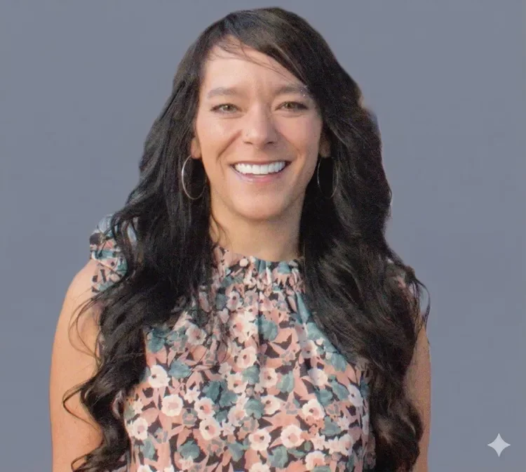 A woman with long black hair, wearing hoop earrings and a floral sleeveless top, smiling against a plain gray background.