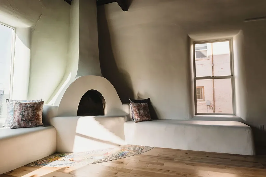 Sunlit room with curved white walls, built-in bench seating with decorative pillows, and a brick building visible through two large windows.