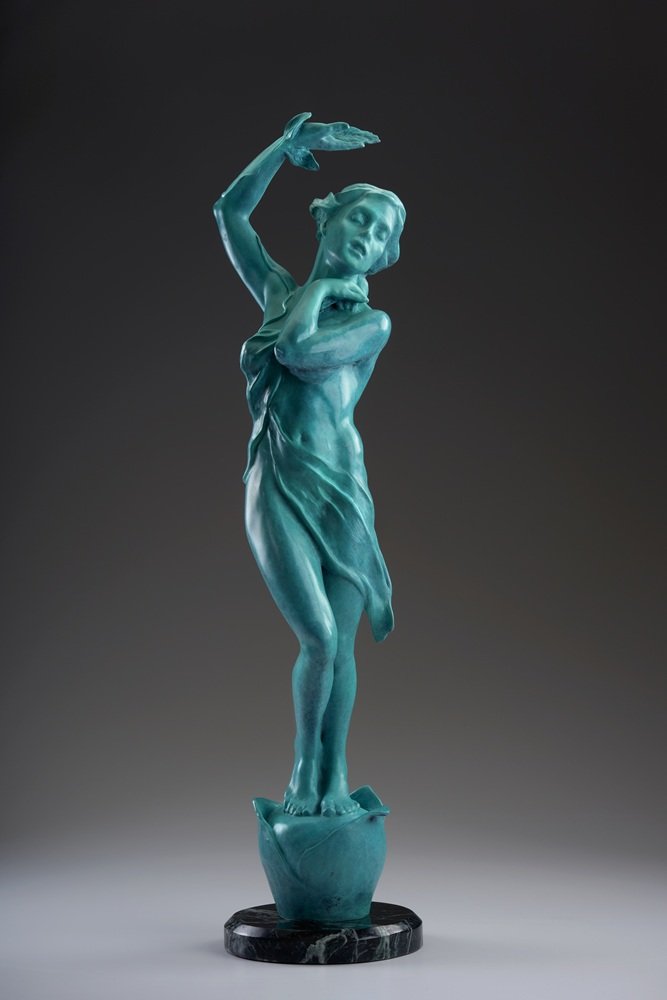 A turquoise-colored statue of a woman standing on a rock, with one arm raised and the other resting on her chest, against a dark background.