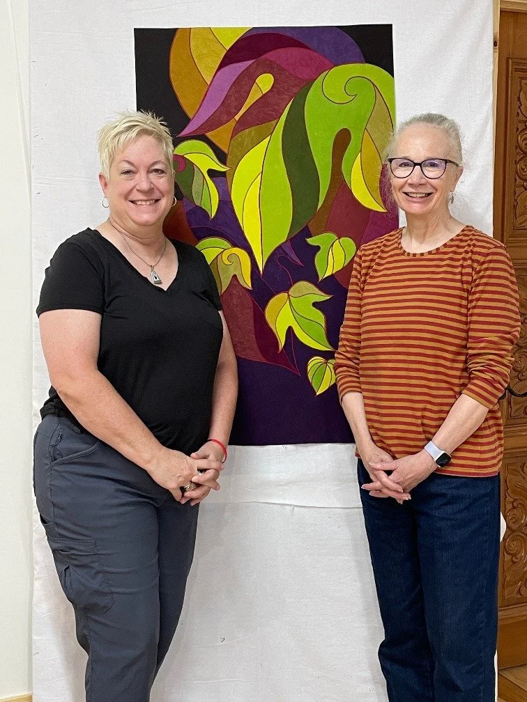 Two women smiling and standing in front of an abstract colorful artwork of swirling green, purple, and yellow leaves on a black background.