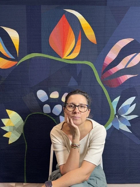 Woman with glasses sitting at a desk, resting her chin on her hand, in front of a colorful floral mural on a dark blue background.