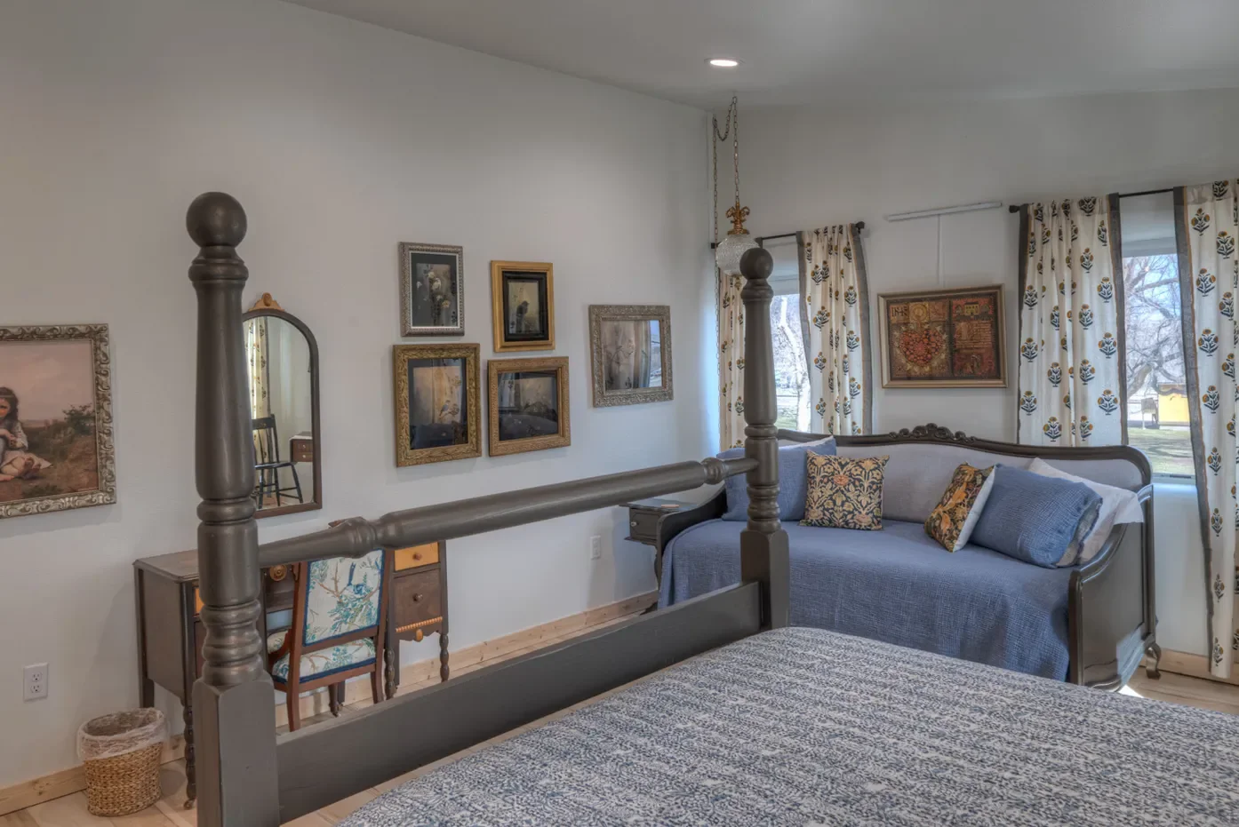 A cozy bedroom with a four-poster bed, a blue daybed with pillows, framed artwork on white walls, a window with patterned curtains, and a small desk with a chair.