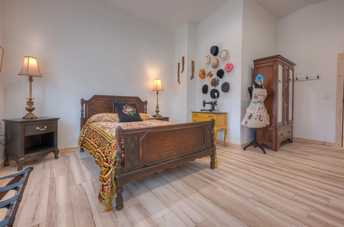 A bedroom with a wooden bed, colorful quilt, black pillow, two side tables with lamps, a yellow cabinet, a vintage sewing machine, a mannequin covered with fabric, and wall dcor including hats and socks on a white wall.