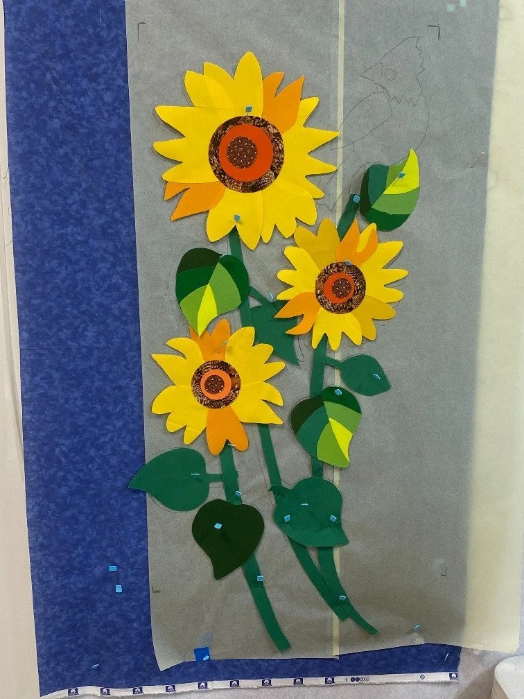 Paper craft of three bright yellow sunflowers with green leaves and stems, attached to a backdrop with a blue and beige background.