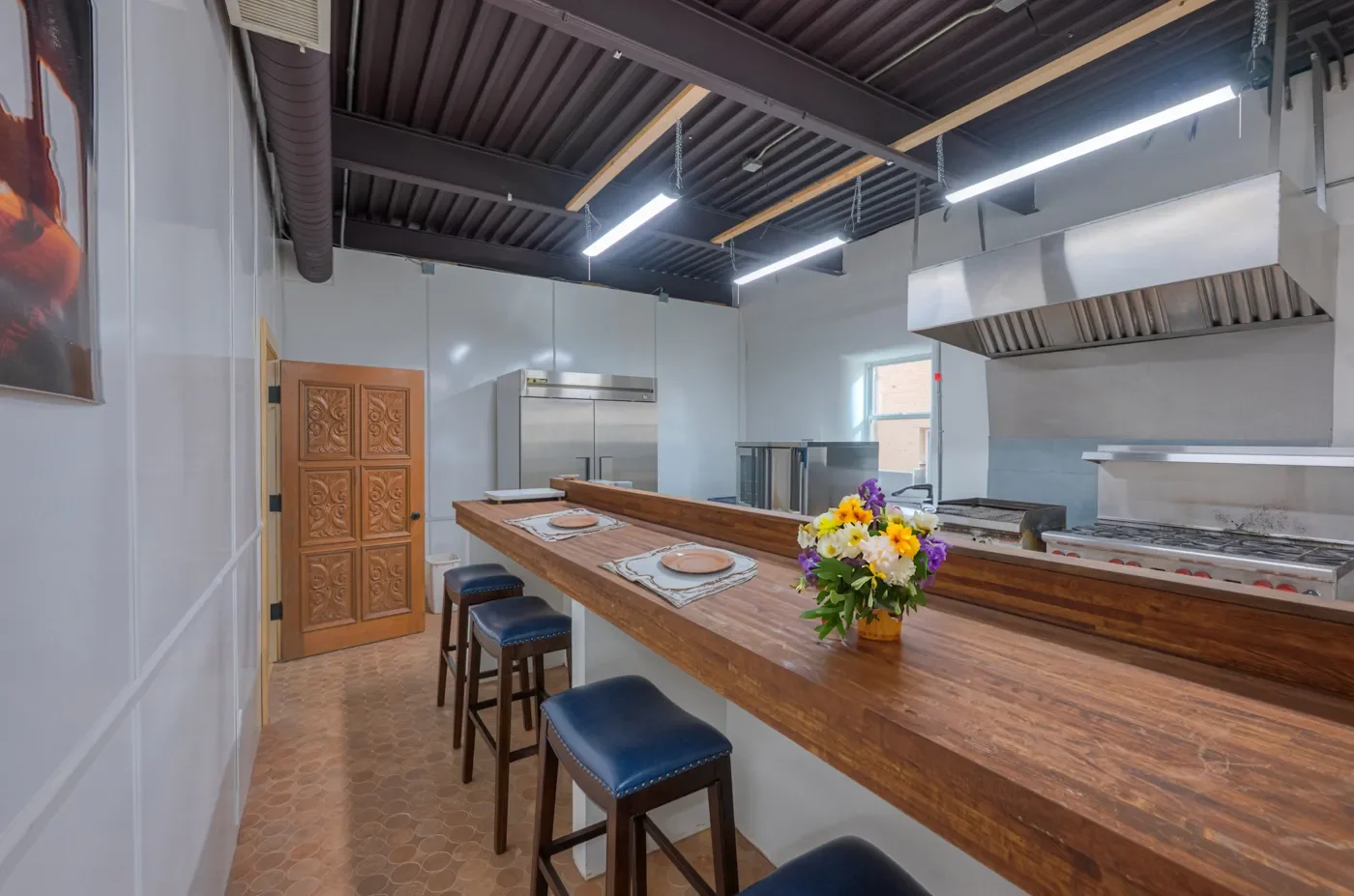 Interior of a modern kitchen with a long wooden counter, bar stools, stainless steel appliances, a stove, and a flower vase with colorful flowers.