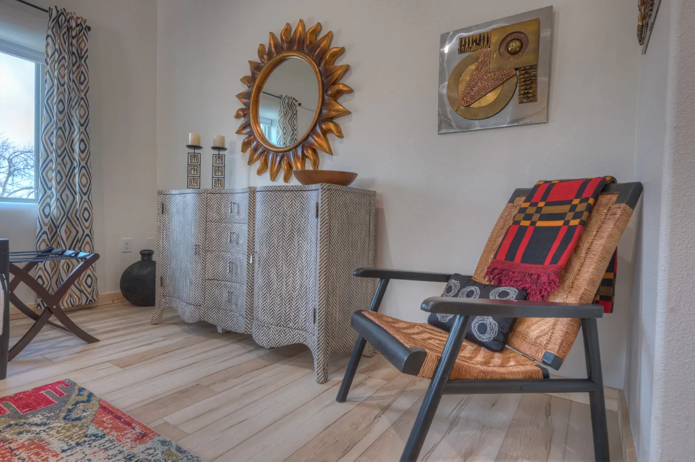 Living room corner with a wooden chair with a woven seat and plaid blanket, a patterned sideboard, a sunburst mirror, and a wall art piece. The room has wood flooring, a window with patterned curtains, and a colorful rug.