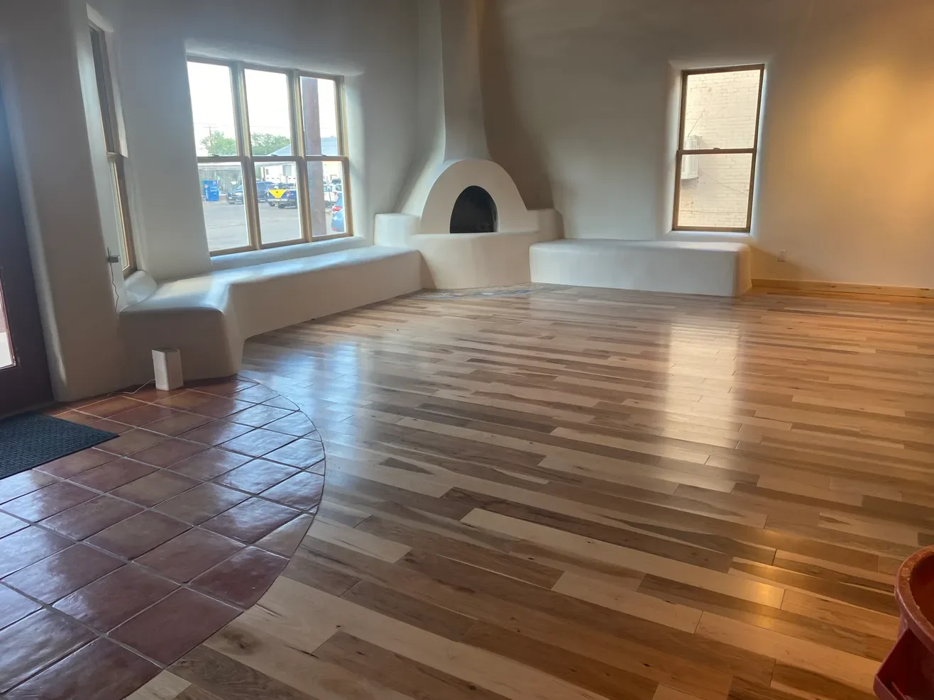 Interior of a room with wooden flooring, a built-in white seating area beneath large windows, a small brick fireplace, and a mix of tile and wood flooring.