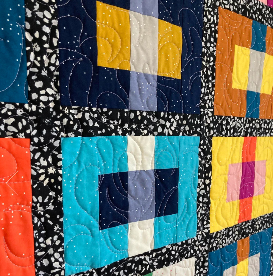 Close-up of a colorful quilt with square patchwork patterns, featuring various colors like blue, yellow, orange, pink, and black with white speckled accents and intricate quilting stitches.