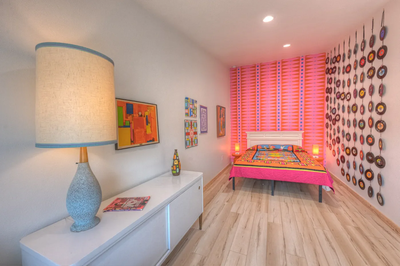 A bedroom decorated with vibrant, colorful, and abstract art, featuring a pink geometric patterned accent wall, a white bed with colorful bedding, and vinyl records hanging on the wall to the right.