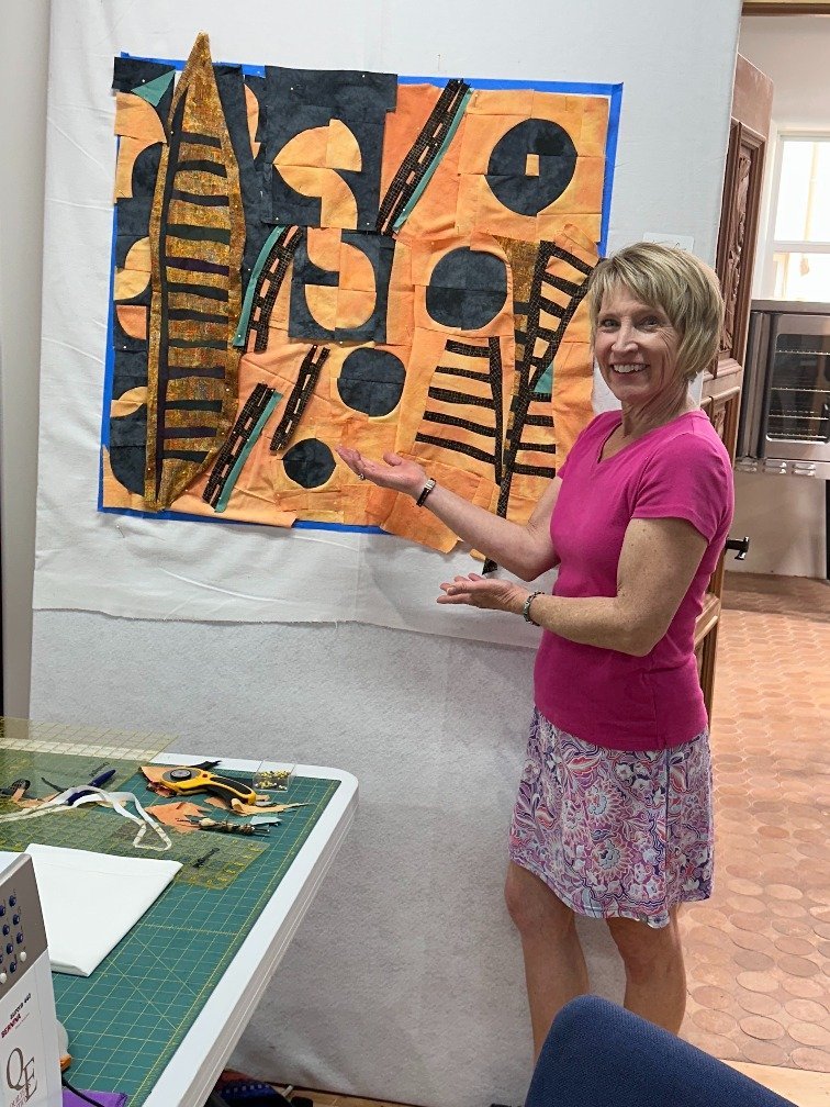 A woman in a pink shirt and floral skirt gesturing towards a quilt on the wall, which features black and orange abstract and geometric shapes. The quilt is displayed on a whiteboard, and sewing tools are on a table nearby.