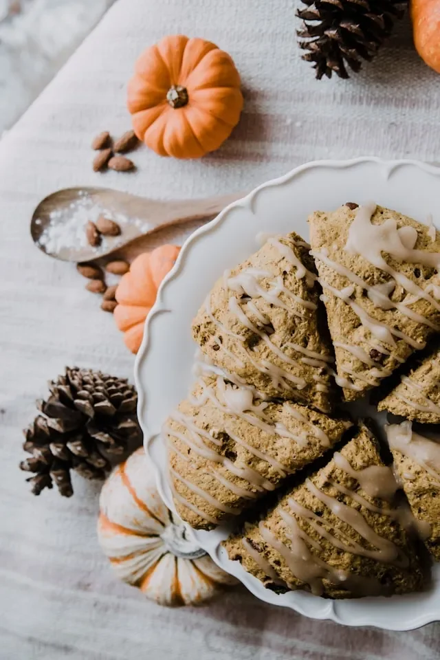 Pumpkin-shaped cookies with white icing drizzled on top, surrounded by small decorative pumpkins and pinecones on a white wooden surface.