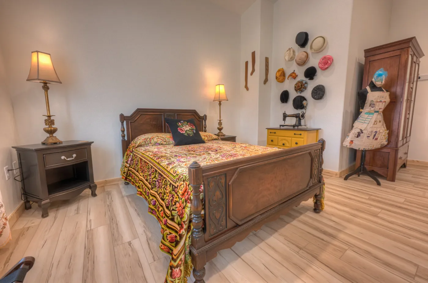 Bedroom with wooden bed, patterned bedspread, floral pillow, two lamps, gray nightstand, yellow dresser, wall decorated with hats and wooden utensils, and a dress form with fabric.