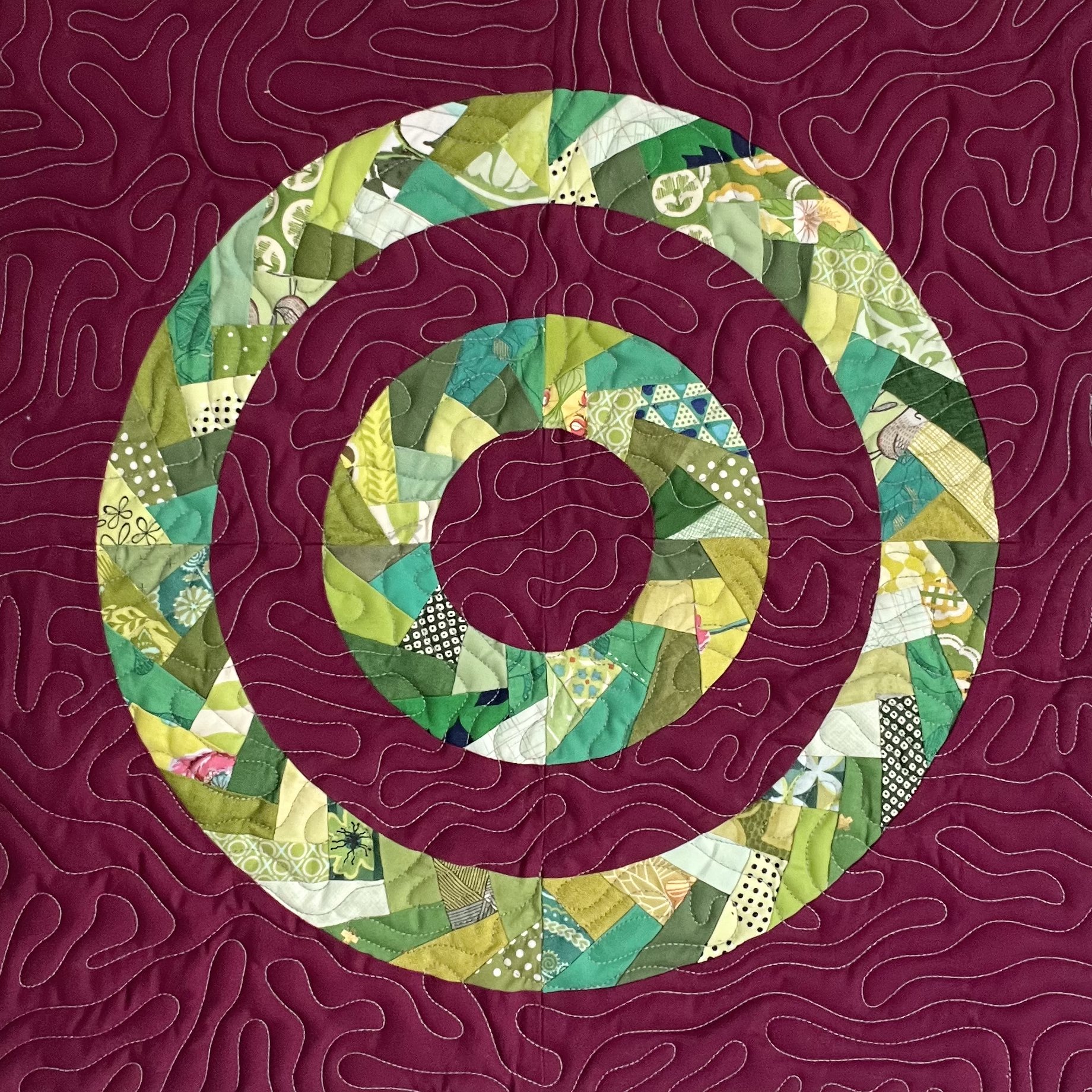 Close-up of a quilted fabric with a question mark design. The background is dark red with stitched wavy lines, and a large question mark shape made of patchwork in various shades of green, yellow, white, and black patterns.