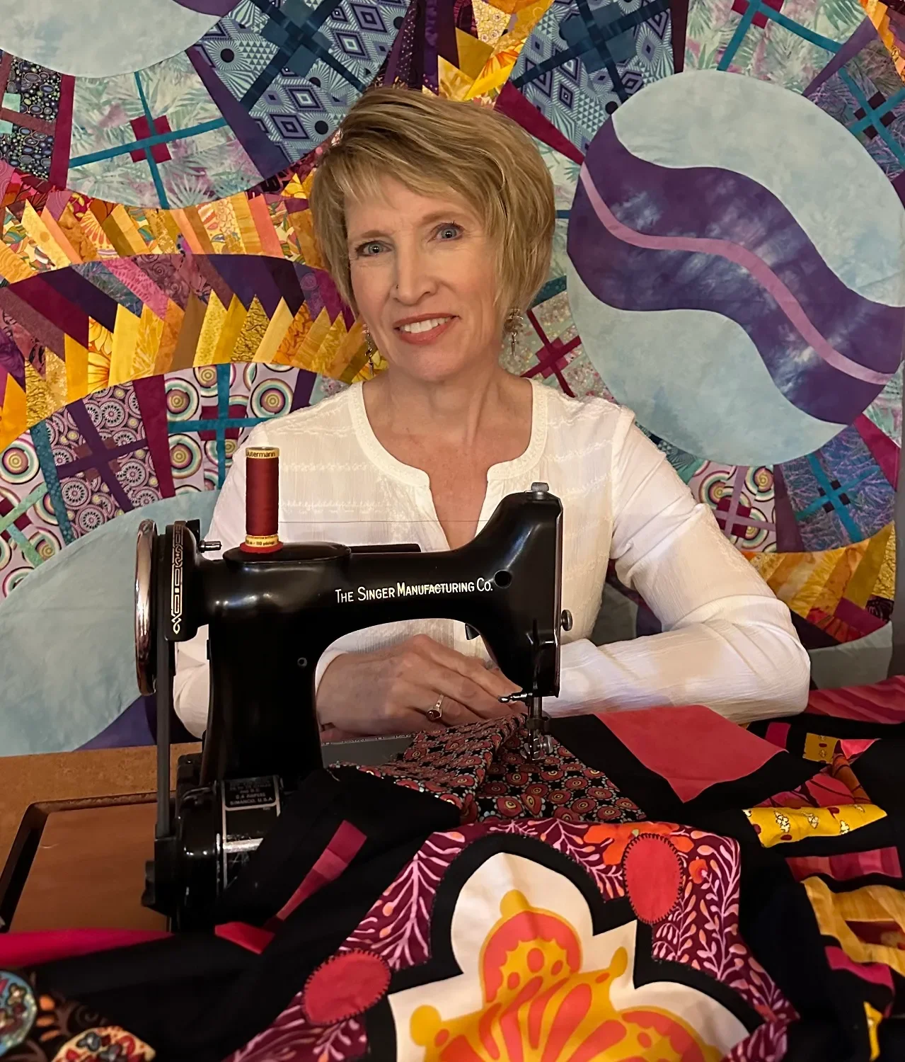 A woman sitting at a table with a sewing machine, surrounded by colorful quilt patterns and fabric, in front of a vibrant quilted backdrop.