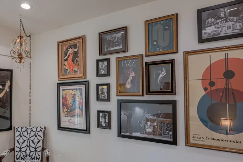 Gallery wall with various framed posters and artwork in a room with a white wall, a chandelier, and a chair with black and white patterned upholstery.