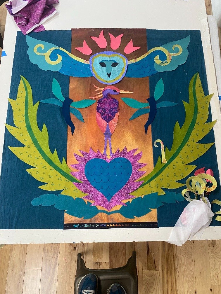 Colorful paper collage artwork featuring a central heart shape, a bird with an elongated neck, and a stylized owl at the top, surrounded by large green leaves, set against a backdrop of warm and cool colors.