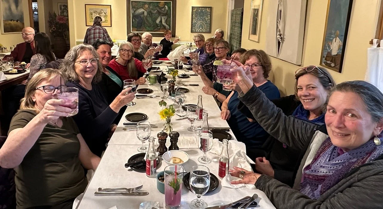 A group of people gathered around a long dinner table celebrating, raising glasses for a toast, with smiles and joyful expressions in a cozy dining room decorated with artwork on the walls.