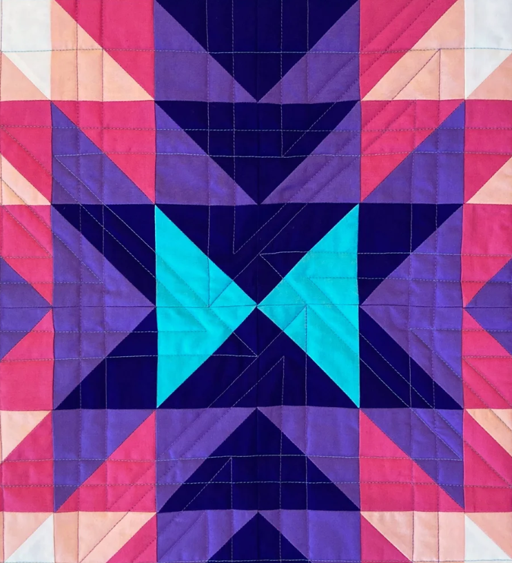 A colorful quilt with geometric patterns in shades of pink, purple, black, and blue arranged symmetrically.