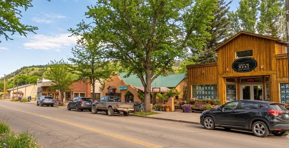 Small town street scene with storefronts, parked cars, trees, and a mountainous background