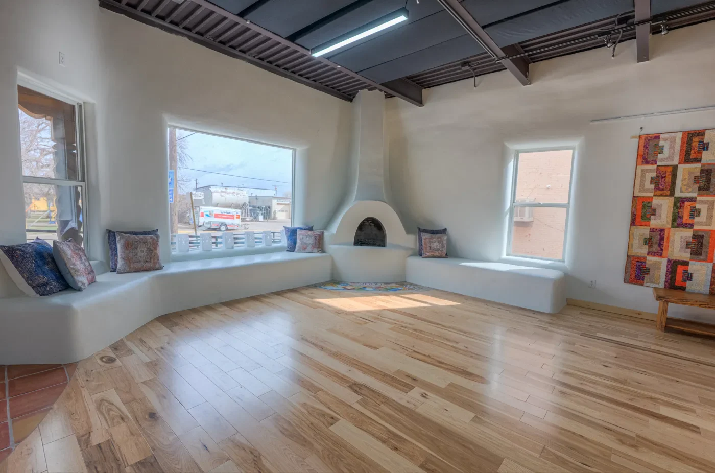 Room with large windows, a built-in seating area with pillows, a fireplace, wooden floor, and a colorful quilt on the wall.