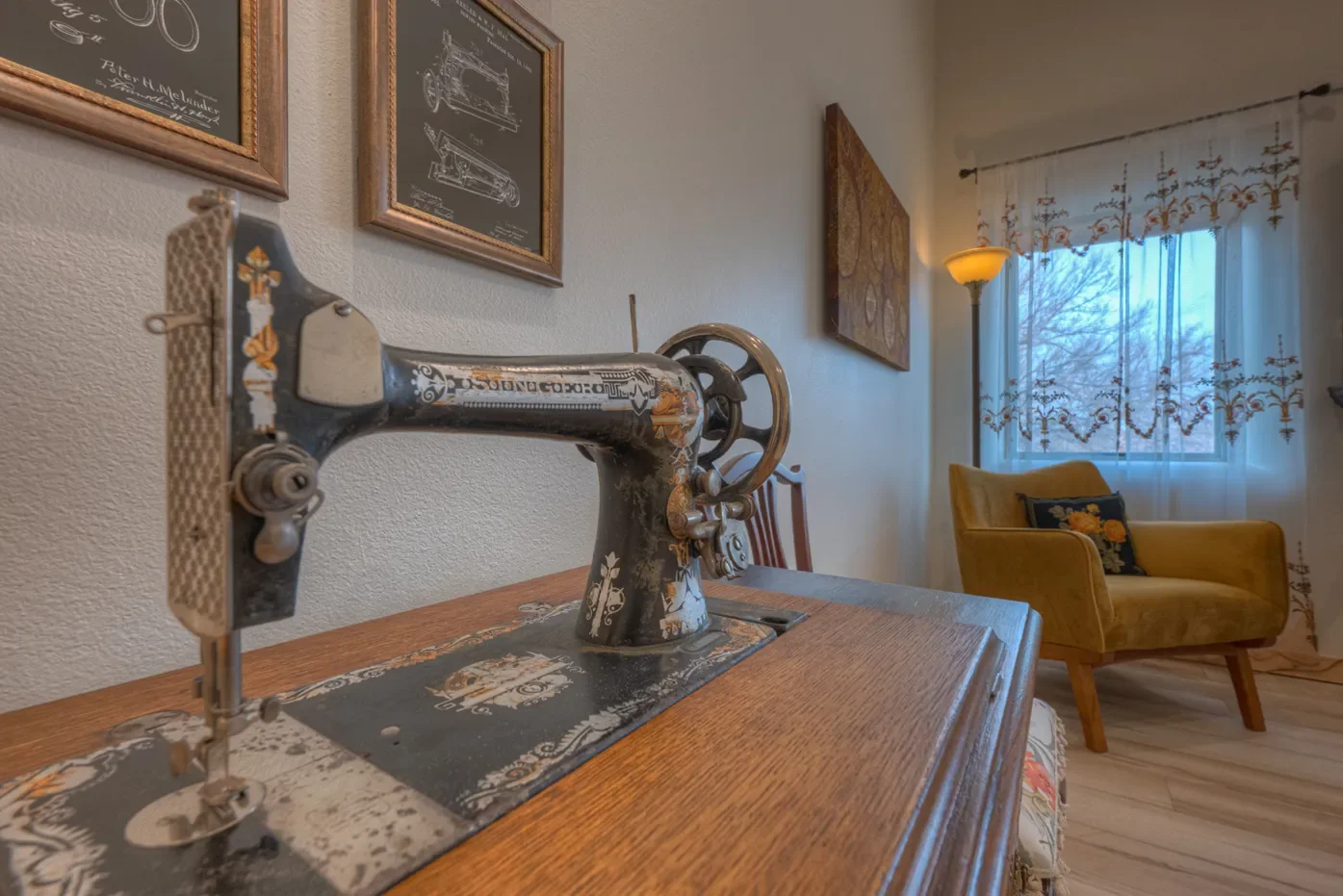 Close-up of a vintage sewing machine on a wooden table in a cozy living room, with framed artwork on the wall, a yellow armchair, and a window with floral curtains in the background.
