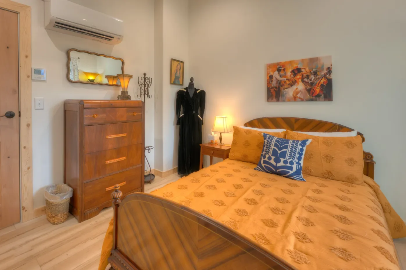 A cozy bedroom with a large wooden bed, orange bedspread, and a blue patterned pillow. There is a wooden nightstand with a lamp, a dresser with a mirror, and a mannequin dressed in black. The room is decorated with framed artwork, a small religious icon, and a mirror. An air conditioning unit is mounted above the dresser.