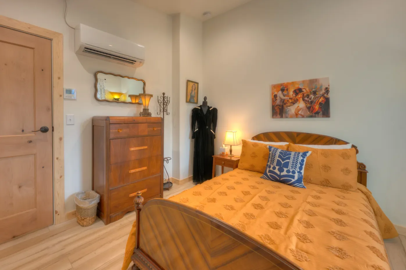 A cozy bedroom with a wooden bed, yellow bedspread and pillows, a nightstand with a lamp, a hanging dress on a stand, a dresser with a mirror, a wall art of jazz musicians, and a framed portrait, with light-colored walls and wooden accents.