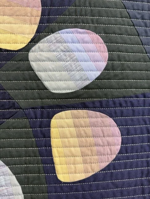 Close-up of a quilt with pastel-colored oval patterns and dark background, stitched with straight lines.