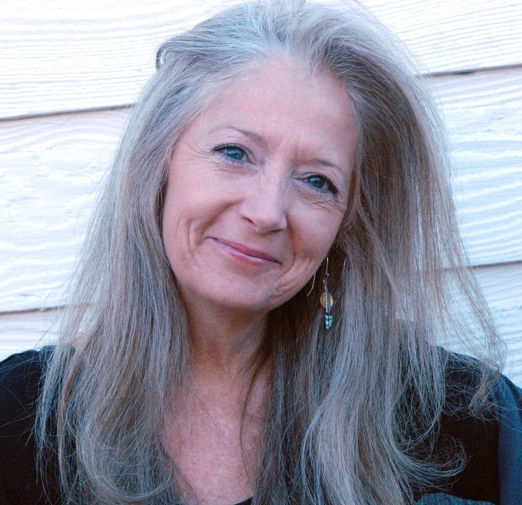 Close-up portrait of a woman with long, gray hair and blue eyes, smiling gently, wearing a black top and earring, with a white wooden wall in the background.