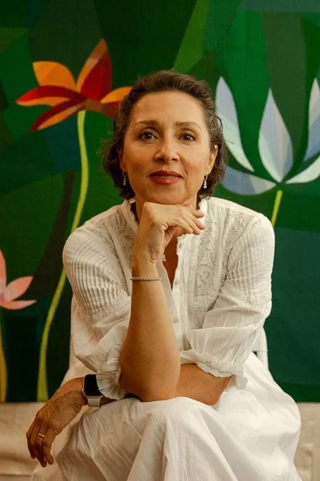 A woman with short brown hair, wearing a white dress, sitting with her chin resting on her hand against a green wall with colorful floral designs.